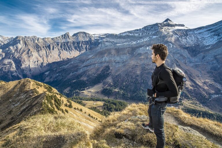 A lone traveler, dressed in hiking gear, is seen navigating a rugged mountain trail. Surrounded by towering peaks and a panoramic view of the expansive landscape, the man is captured in mid-motion, symbolizing adventure and exploration. The clear blue sky above contrasts with the earthy tones of the rocky terrain, highlighting the vastness and beauty of nature. His focused expression and determined stride suggest a journey filled with both challenge and exhilaration